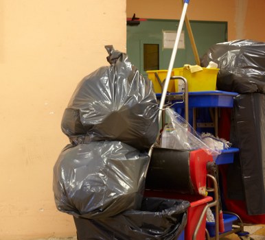 The image depicts a collection of garbage bags and waste containers positioned against a light yellow wall, likely within an indoor or sheltered area such as a storage room or back area of a commercial property. Several large, black plastic rubbish bags are stacked on top of each other, with some leaning against a small red trolley or cart. The bags are filled with waste, their surfaces appearing crinkled and shiny, indicating they are tightly packed. To the right, a larger black waste bag is also visible, resting on a slightly elevated surface, possibly a shelf or table. Behind the bags, a blue container with a lid and a yellow container are partially visible, suggesting additional waste management supplies. The background includes a closed door painted in a darker green shade, with some minimal signage or notices attached to it. The environment is utilitarian, with a focus on waste storage, typical for a business involved in rubbish collection or clearance services in the Brimsdown area. This scene illustrates the type of waste collection setup that Commercial Waste Brimsdown might handle, emphasizing local commercial rubbish accumulation and disposal.