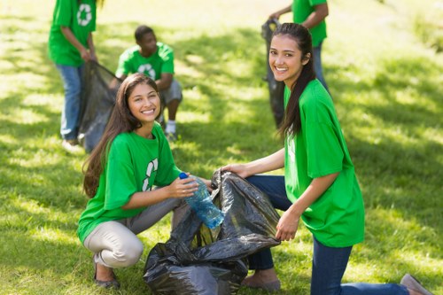 Four individuals participating in a community clean-up event outdoors on a grassy area, wearing bright green T-shirts featuring a white recycling symbol on the front. In the foreground, two young women are smiling at the camera while holding a large black rubbish bag filled with waste, with one woman kneeling on the grass and the other standing. The woman kneeling has long brown hair and is holding a clear plastic bottle, ready to place it into the bag, while the standing woman has long dark hair pulled back. In the background, two more volunteers are visible: one holding the bag open and the other standing nearby, both also in green T-shirts, engaged in collecting litter. The setting appears to be a park or a green open space with sunlight filtering through trees, creating a lively atmosphere of environmental community effort, aligned with waste management and rubbish collection activities in the local area.
