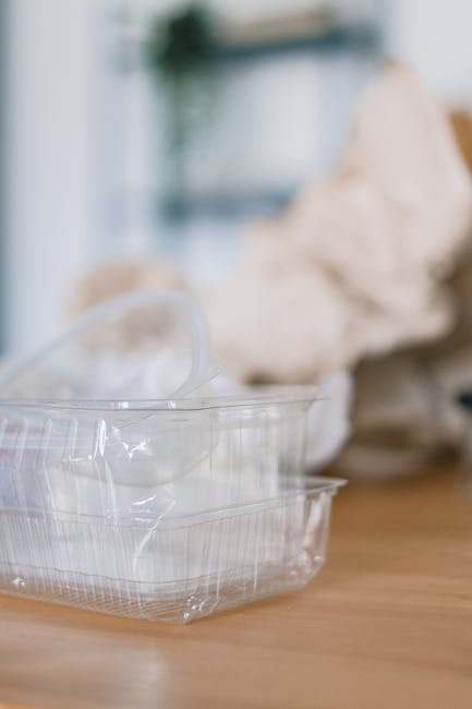The image shows a person placing a transparent plastic container into a white recycling bin labeled 'PLASTIC' on a wooden table. The person is wearing a beige pair of trousers and an orange top, with