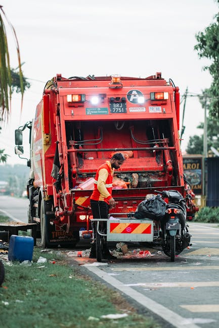 A waste collection worker dressed in a high-visibility yellow and red uniform is performing rubbish removal on a street, standing beside a large, red garbage truck with its rear loading mechanism visi