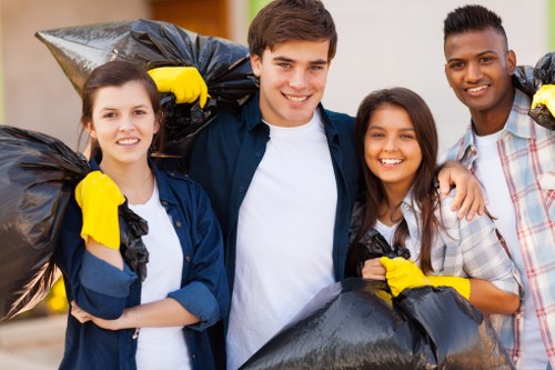 In this image, four young adults stand outdoors, each holding large black rubbish bags filled with waste materials. The individuals are smiling and appear to be engaged in a community or group clean-up activity. They are dressed casually, with some wearing yellow protective gloves for handling rubbish, and their clothing includes denim, patterned shirts, and casual tops. The background suggests an urban or suburban environment with blurred greenery and buildings. The scene is well-lit, indicating daytime, and the overall atmosphere is positive and cooperative. This visual relates to waste collection or rubbish removal efforts typically supported by services such as those offered by Commercial Waste Brimsdown, serving the Brimsdown area, which is part of the London postal code region. The image exemplifies a community initiative to clear rubbish from public space, aligning with the concept of effective commercial waste solutions in the local area.